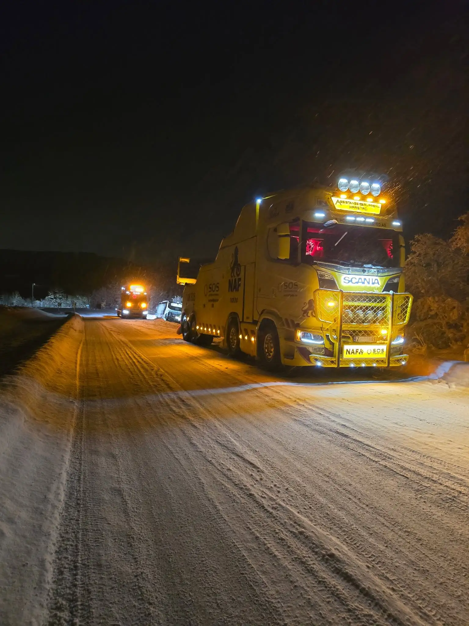 Gul bergingsbil med arbeidslysene tent på en snødekt vei om natten